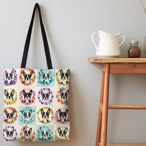 K&ouml;nnte beinhalten: Eine Tragetasche mit einem sich wiederholenden Muster von Boston Terrier-Portr&auml;ts, jedes von einem Blumenkranz umrahmt. Die Tasche hat einen cremefarbenen Hintergrund, schwarze Griffe und wird vor einer hellgrauen Wand pr&auml;sentiert.