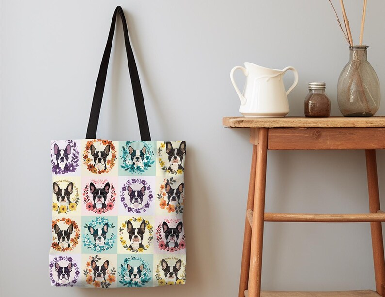 K&ouml;nnte beinhalten: Eine Tragetasche mit einem sich wiederholenden Muster von Boston Terrier-Portr&auml;ts, jedes von einem Blumenkranz umrahmt. Die Tasche hat einen cremefarbenen Hintergrund, schwarze Griffe und wird vor einer hellgrauen Wand pr&auml;sentiert.