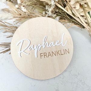 Pode incluir: Um sinal redondo de madeira com o nome "Raphael Franklin" escrito em letras brancas em relevo.