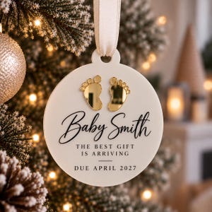 Puede incluir: Un adorno redondo blanco con una cinta color crema, que presenta dos huellas de bebé en acabado espejo dorado. El texto dice Baby Smith, THE BEST GIFT IS ARRIVING y DUE APRIL 2027 en letras negras, colgado de una rama de árbol de Navidad.
