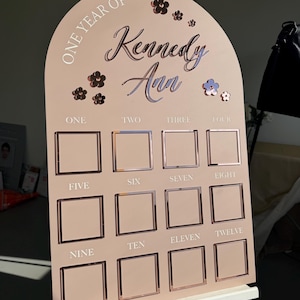 K&ouml;nnte beinhalten: Eine rosafarbene Meilensteintafel mit dem Text "ONE YEAR OF Kennedy Ann" und den Zahlen eins bis zw&ouml;lf. Die Tafel hat quadratische Rahmen f&uuml;r Fotos und Blumendekorationen. Sie wird auf einer wei&szlig;en Staffelei pr&auml;sentiert, ideal f&uuml;r den ersten Geburtstag.