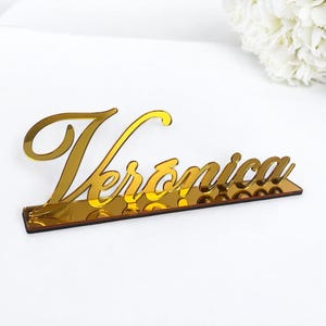 May include: Gold mirror acrylic table number with the name Veronica in a cursive font.