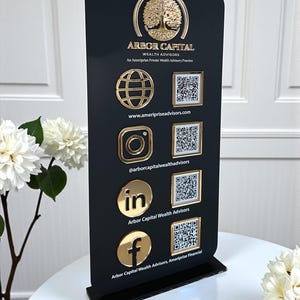 Könnte beinhalten: Ein schwarzer Displayständer mit dem Logo von Arbor Capital Wealth Advisors und Kontaktinformationen. Goldfarbene Logos für Globus, Instagram, LinkedIn und Facebook werden zusammen mit QR-Codes angezeigt. Der Ständer steht auf einem weißen Tisch mit Blumen.