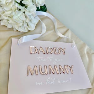 Puede incluir: Un cartel rosa con las palabras "Daddy Time to give Mummy our last name!" en letras doradas.