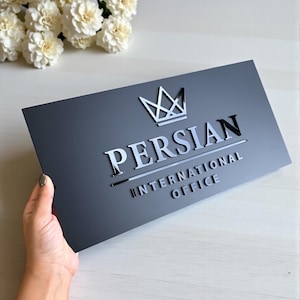 Może przedstawiać: Prostokątny czarny znak z napisem "PERSIAN INTERNATIONAL OFFICE" w srebrnych literach. Srebrny wzór korony znajduje się nad słowem "PERSIAN". Znak jest trzymany w dłoni, z białymi kwiatami w tle.