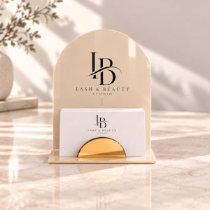 Può includere: Un porta biglietti da visita in acrilico beige a forma di arco con il logo nero e la scritta "LB LASH & BEAUTY STUDIO". Una pila di biglietti da visita bianchi con lo stesso logo nero è tenuta ferma da un piccolo elemento in metallo dorato.