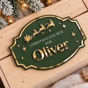 May include: A rectangular wooden box featuring a decorative dark green plaque with gold-coloured accents. The plaque displays a silhouette of Santa in a sleigh with reindeer, stars, and the text "CHRISTMAS EVE BOX FOR Oliver" in gold lettering.