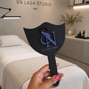 Op de afbeelding: Een zwarte handspiegel met een gebogen bovenrand, voorzien van een verhoogd, glanzend zwart "SN" monogramlogo en de tekst "SN LASH STUDIO" in het wit gedrukt. De spiegel heeft een lange, rechte steel met een klein rond gat aan het uiteinde.