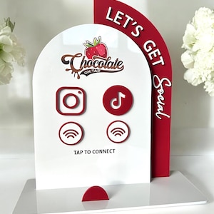 Op de afbeelding: Een wit bord met rode accenten en de tekst "Let's Get Social" aan de rechterkant. Het bord heeft ook een logo voor "Chocolate on Tap" met een aardbeienafbeelding. Het bord heeft pictogrammen voor Instagram, TikTok en Wi-Fi.