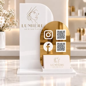 May include: A white and gold acrylic sign featuring the text LUMIÈRE AESTHETICS, a line-art face logo, Instagram and Facebook icons, and two QR codes. A matching white business card with the same logo sits in a small holder on the base.