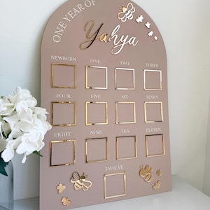 Puede incluir: Un tablero de hitos arqueado de color topo con detalles dorados. El tablero presenta el texto "ONE YEAR OF Yahya" y doce marcos cuadrados para fotos. Adornos de mariposas doradas.