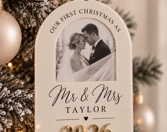 May include: An arched cream-colored acrylic Christmas ornament featuring a black and white photograph of a couple. The text reads OUR FIRST CHRISTMAS AS Mr & Mrs TAYLOR 2026, with the year 2026 displayed in raised gold-toned metallic numbers.