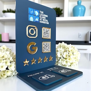 Pode incluir: Um expositor azul marinho com detalhes dourados. O expositor apresenta os logotipos do Instagram e Google, códigos QR e cinco estrelas douradas. O texto no expositor diz "The Leading Clinic at Royal Kingston", "Follow Us" e "Leave Us a Review".