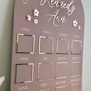 K&ouml;nnte beinhalten: Eine mauvefarbene Meilensteintafel aus Holz mit dem Text "ONE YEAR OF Kennedy Ann" und Blumenakzenten. Sie verf&uuml;gt &uuml;ber zw&ouml;lf quadratische Fotorahmen mit den Zahlen eins bis zw&ouml;lf. Ein dekoratives St&uuml;ck f&uuml;r das erste Lebensjahr des Babys.