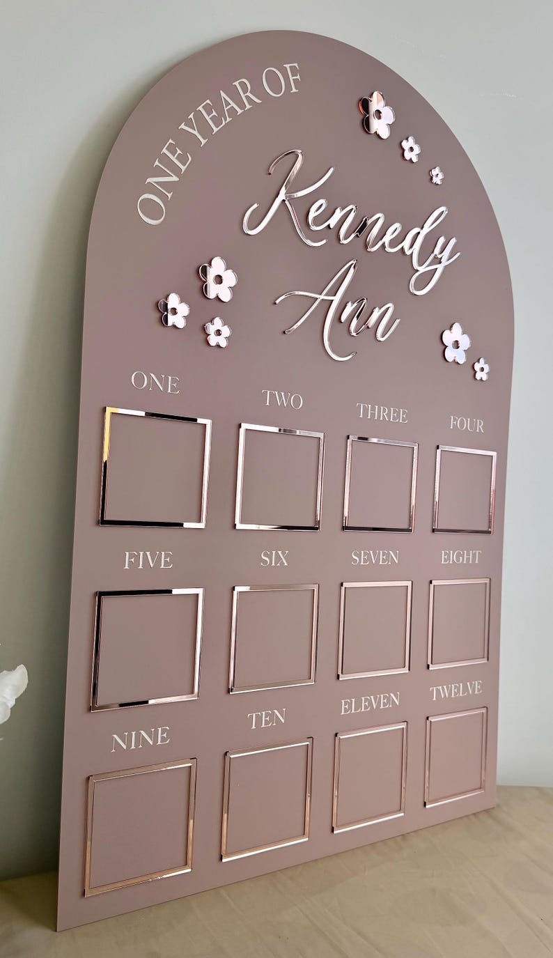K&ouml;nnte beinhalten: Eine mauvefarbene Meilensteintafel aus Holz mit dem Text "ONE YEAR OF Kennedy Ann" und Blumenakzenten. Sie verf&uuml;gt &uuml;ber zw&ouml;lf quadratische Fotorahmen mit den Zahlen eins bis zw&ouml;lf. Ein dekoratives St&uuml;ck f&uuml;r das erste Lebensjahr des Babys.