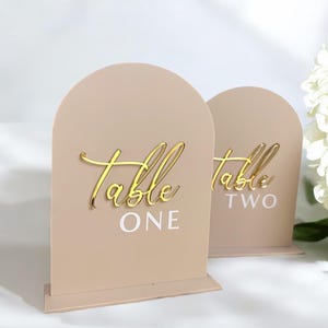 May include: Two light brown acrylic table number signs with a rounded arch top. The signs have a gold glitter finish and the text "Table One" and "Table Two" in white and gold lettering.