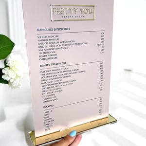 Pode incluir: Um menu de salão rosa e dourado com o texto "PRETTY YOU BEAUTY SALON" em letras douradas. O menu lista serviços de manicure, pedicure, tratamentos de beleza e depilação. O menu é segurado por uma mão com manicure azul claro.