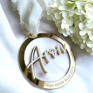 Pode incluir: Um ornamento redondo dourado com uma inscrição branca que diz "Aron" e "Mijn eerste kerst 2024".
