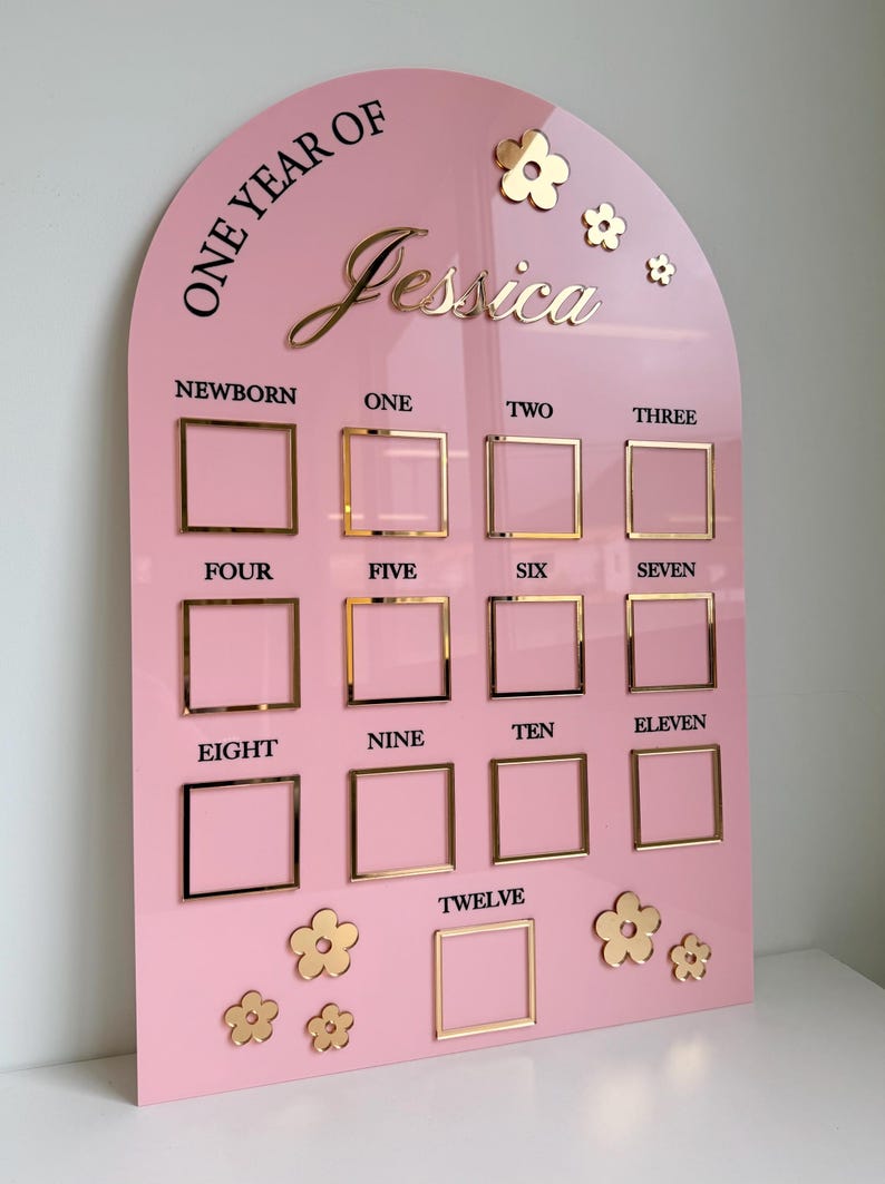 K&ouml;nnte beinhalten: Rosafarbene Meilensteintafel aus Acryl mit goldenen Akzenten. Enth&auml;lt den Text "One Year of Jessica" und Rahmen f&uuml;r jeden Monat. Inklusive goldener Blumenverzierungen. Ein dekoratives St&uuml;ck f&uuml;r Babyfotos.