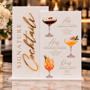 Könnte beinhalten: Ein weißes Acrylschild mit dem goldenen Schriftzug Signature Cocktails und einer Menüliste für His Espresso Martini, Hers Passionfruit Martini und Ours Aperol Spritz, inklusive Abbildungen der Getränke, Zutaten und goldener Schrift.