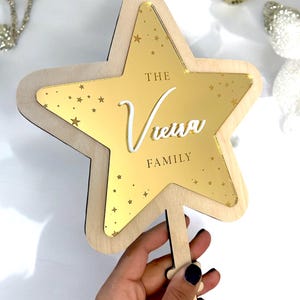 Könnte beinhalten: Ein sternförmiger Holzschmuck mit einem goldenen Spiegeleinsatz. Der Schmuck hat einen schlüsselförmigen Griff und trägt den Text "The Vienna Family" in weißen Buchstaben.