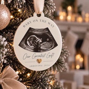 May include: A round white ceramic ornament hanging from a beige ribbon. The ornament features a black and white ultrasound image, the text "BABY ON THE WAY", "Our Greatest Gift", a small gold heart icon, and the text "ARRIVING 2026" printed on the front.