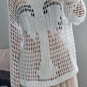 Könnte beinhalten: Weißer Häkelpullover mit lockerer Passform und einem großen, offenen Maschenmuster. Der Pullover ist durchsichtig und hat einen runden Ausschnitt.
