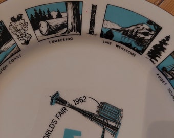May include: A white ceramic plate with a blue and black design depicting scenes from the 1962 Seattle World's Fair. The plate features illustrations of lumbering, Lake Wenatchee, Puget Sound, and the Space Needle. The text "1962 Seattle World's Fair" is printed on the plate.