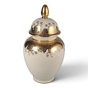 May include: A cream-colored ceramic ginger jar with a removable lid featuring a gold finial. The jar and lid are decorated with a metallic gold lace-like pattern and small floral motifs. The base and rim of the lid are also accented with gold.