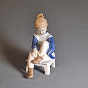 以下が含まれることがあります： 小さな白いスツールに座り、青と白の服を着た若い人の磁器製の置物。置物は茶色の靴を履いています。髪はお団子にまとめられています。