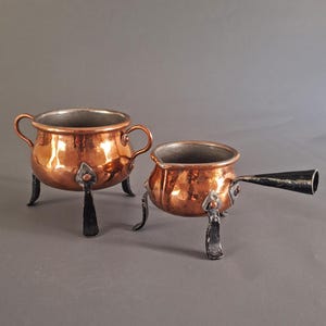 Könnte beinhalten: Zwei Kupferkessel mit silbernen Innenräumen und schwarzen Metallbeinen. Ein Kessel hat zwei Griffe, der andere einen langen Griff. Die Kessel stehen auf grauem Hintergrund. Das Kupfer hat eine glänzende, reflektierende Oberfläche.