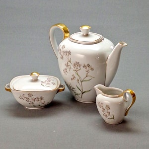 May include: A white porcelain tea set with a floral design. The set includes a teapot, creamer and sugar bowl. The teapot has a gold handle and spout. The creamer and sugar bowl have gold accents.