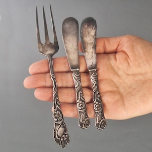 Puede incluir: Un juego de cubiertos antiguos, que incluye un tenedor y dos cuchillos de mantequilla, sostenidos en una mano. La cubertería tiene detalles florales y de volutas ornamentados en los mangos. El metal tiene un aspecto vintage deslustrado.