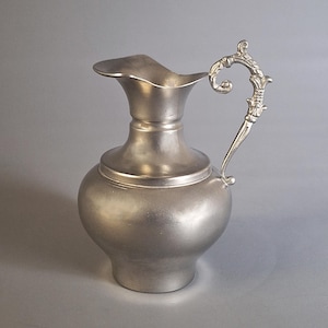 Może przedstawiać: Srebrny dzban z ozdobnym uchwytem. Dzban ma pękaty korpus i rozszerzający się dzióbek. Uchwyt jest zdobiony wzorem zwojów. Dzban prawdopodobnie wykonany jest z cyny lub podobnego metalu.