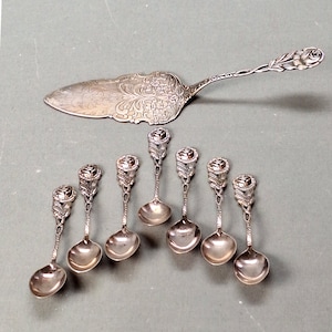 Może przedstawiać: Zestaw ośmiu srebrnych łyżeczek do herbaty z uchwytami w kształcie pączka róży i srebrny nóż do ciasta z uchwytem w kształcie pączka róży i ozdobnym wzorem.