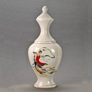 May include: White ceramic vase with a lid, featuring a painted design of a woman in a red and white dress with flowing blue and green ribbons.