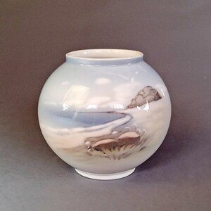 Può includere: Un vaso rotondo in ceramica azzurra con un dipinto paesaggistico. Il dipinto raffigura una scena di spiaggia con acqua, nuvole e una piccola collina. Il vaso ha una base bianca e un bordo leggermente svasato.
