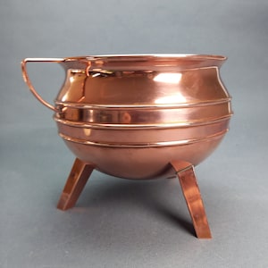 Op de afbeelding: Een kleine koperen pot met drie poten. De pot heeft een rond lichaam en een enkele handgreep.