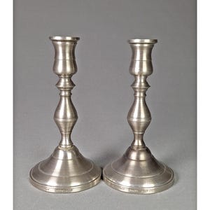 May include: Two silver-colored candlesticks with a classic design. Each candlestick has a wide, circular base, a slender, elongated body with decorative curves, and a top designed to hold a candle. The candlesticks are made of metal and have a matte finish.