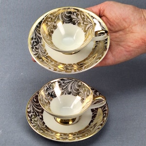 May include: Two white teacups and saucers with gold leaf designs. The cups and saucers have a delicate floral pattern.