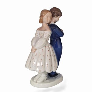Könnte beinhalten: Eine Porzellanfigur, die zwei Figuren zeigt, die Rücken an Rücken auf einem ovalen Sockel stehen. Eine Figur trägt ein weißes schulterfreies Kleid mit braunem Blumenmuster, die andere einen dunkelblauen Anzug mit weißem Kragen.