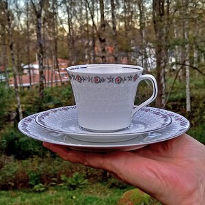 以下が含まれることがあります： ピンクとグレーの色合いの花柄が施された、白いティーカップとソーサーのセット。カップは繊細なエンボス加工が施され、金色の縁取りがされています。