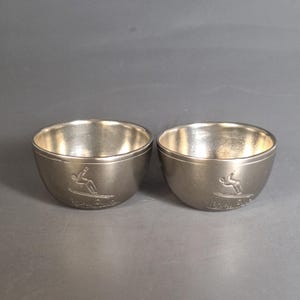 May include: Two silver metal cups with a skier design on the bottom. The cups are about 2 inches tall and 2 inches wide.