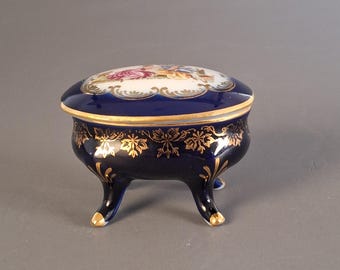 May include: A cobalt blue porcelain trinket box with a lid featuring a painted scene. The box has three feet and is adorned with gold leaf accents. The lid and base are trimmed with gold. The box is approximately 4 inches in diameter.