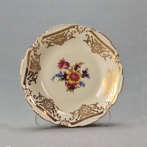 May include: A white ceramic bowl with a gold rim and floral design. The bowl has a scalloped edge and is decorated with a floral pattern in shades of pink, yellow, and purple.