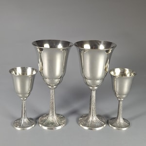 Könnte beinhalten: Vier silberne Kelche in verschiedenen Größen werden vor einem grauen Hintergrund präsentiert. Die Kelche haben eine tulpenförmige Schale, einen schlanken Stiel und einen strukturierten, ausgestellten Fuß. Das Metall hat eine polierte, reflektierende Oberfläche.