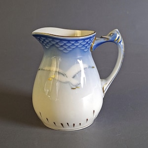 May include: A white and blue ceramic pitcher with a swan design. The pitcher has a blue scaled pattern at the top, a swan-shaped handle, and gold accents. The pitcher is approximately 7 inches tall.