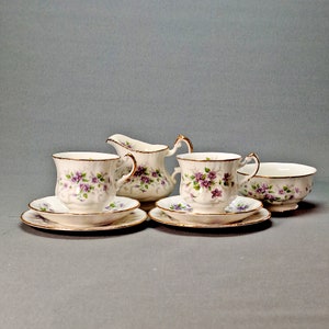 Könnte beinhalten: Ein Set aus vier weißen Porzellan-Teetassen und Untertassen mit einem floralen Muster aus lila und weißen Blumen. Die Tassen und Untertassen haben einen goldenen Rand.
