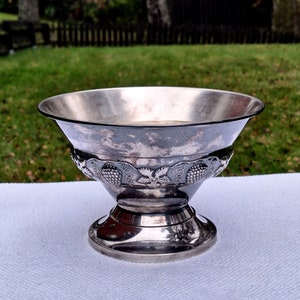 May include: A silver bowl with a decorative grape and leaf design around the rim. The bowl is on a pedestal base.
