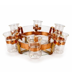 Könnte beinhalten: Ein Vintage-Servierset aus Glas und Kupfer. Das Set besteht aus einer großen, klaren Glasschale, die von einem Kupferband umgeben ist, und sechs kleinen Schnapsgläsern. Die Schnapsgläser werden von Kupferhalterungen gehalten. Das Kupfer hat eine warme, gealterte Patina.
